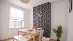 A dining area with a wooden table and chairs, a patterned wall, a window with blinds, and a potted plant. - Modern 3BR Home with Free Parking & Fast Wi-Fi (Rugby)