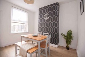 A dining area with a wooden table and chairs, a patterned wall, a window with blinds, and a potted plant. - Modern 3BR Home with Free Parking & Fast Wi-Fi (Rugby)