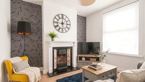 A living room with a fireplace, a clock, a television, and a sofa. - Modern 3BR Home with Free Parking & Fast Wi-Fi (Rugby)