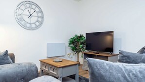 Un salon moderne avec une grande horloge murale, un téléviseur à écran plat et des meubles recouverts d’un tissu gris.