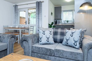 Een moderne woonkamer met een grijze bank, twee gekleurde kussens, een houten salontafel, en een eethoek met een witte tafel en stoelen.