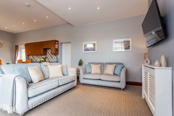 A living room with two sofas, a television mounted on the wall, and a kitchen in the background.