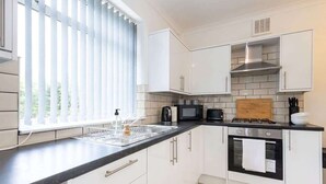 A kitchen with white cabinets, a black countertop, a window with blinds, a microwave, a toaster, and an oven. - Bright & Spacious 3BED Home w Garden in St Helens (St Helens)