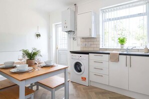 A modern kitchen with a built-in washing machine, a dining table set with plates and glasses, and a window with a view of greenery. - Spacious 3BR House with Garden in St Helens (St Helens)