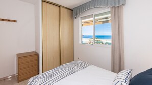 A bedroom with a bed, a wooden wardrobe, a small cabinet, and a window with a view of the sea. - Mar Internum: Family Apartment in La Pineda (La Pineda)