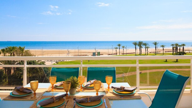 A dining table set with plates, glasses, and napkins, overlooking a beach with palm trees and a clear sky. - Mar Internum: Family Apartment in La Pineda (La Pineda)