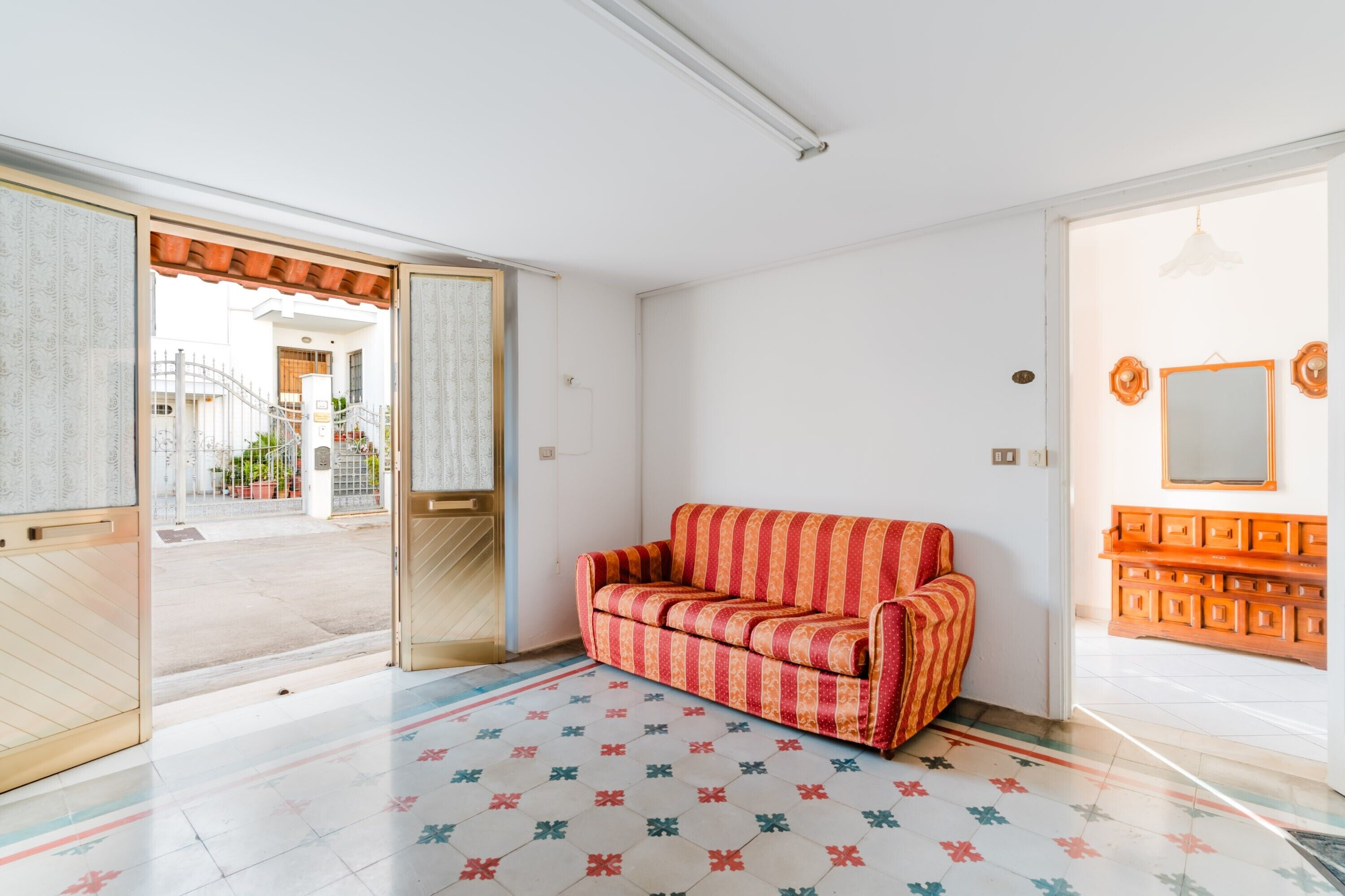 An open room with a patterned floor, a red-striped sofa, a wooden cabinet, and a door leading outside.
