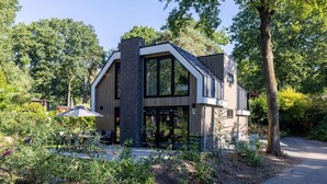 A modern two-story house with a large glass facade, a brick chimney, and a patio with outdoor seating. - Stunning private villa for 10 guests with WIFI, pool, TV and terrace (Apeldoorn)
