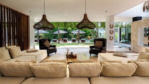 Amplia sala de estar con un gran sofá, dos sillones y una mesa de centro. Se observan dos lámparas colgantes de mimbre y una pared de acento de piedra. La habitación tiene ventanas amplias que dan a una piscina y un área de estar al aire libre.