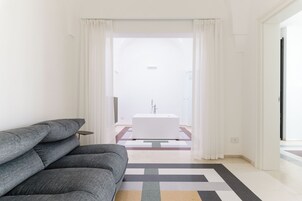 Una habitación de hotel moderna con un sofá gris, una pequeña mesa redonda y vistas a un baño con bañera.