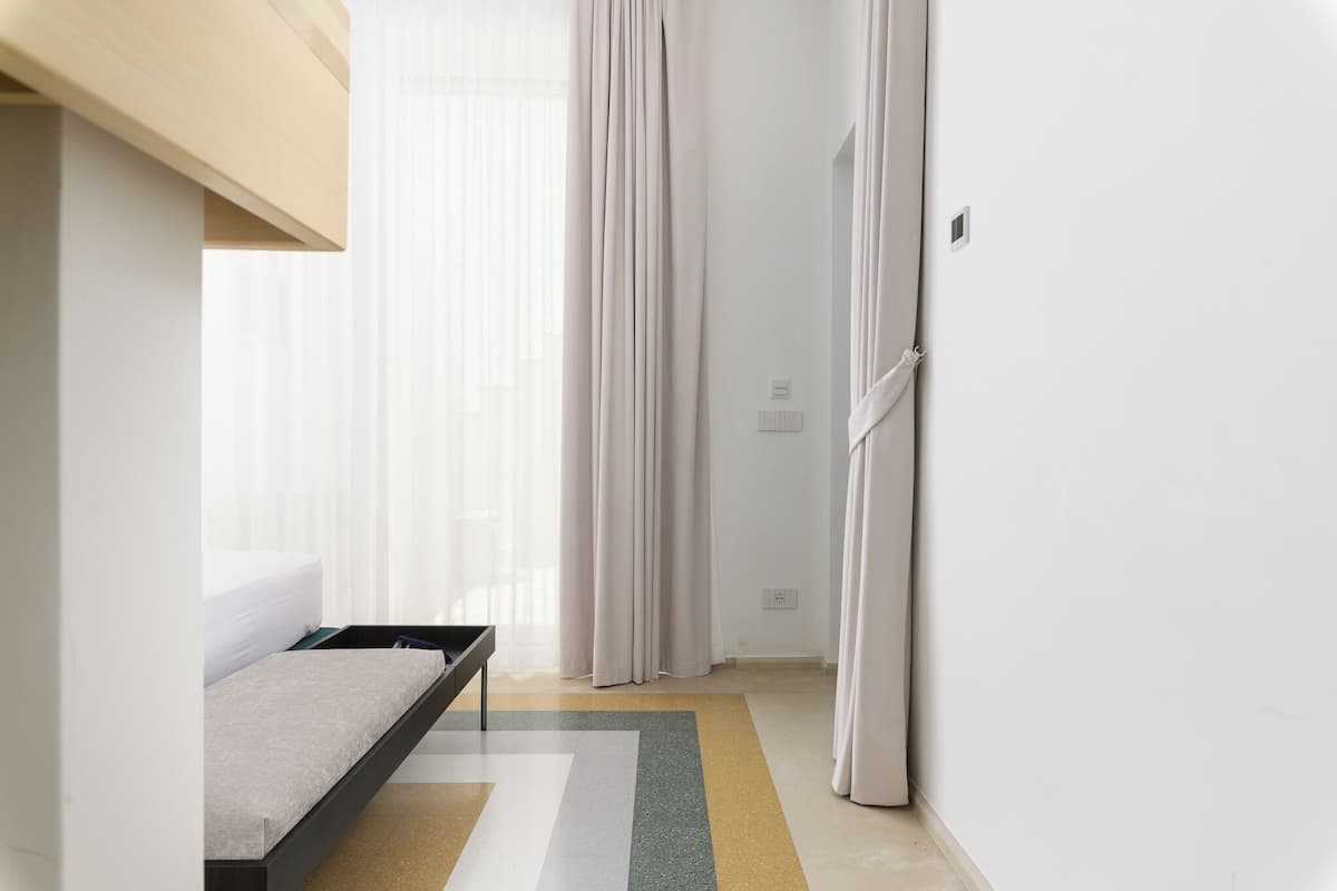 Ein modernes Hotelzimmer mit Bett, einem kleinen Tisch, einem Fenster mit leichtem Vorhang und einer Tür mit Vorhang.