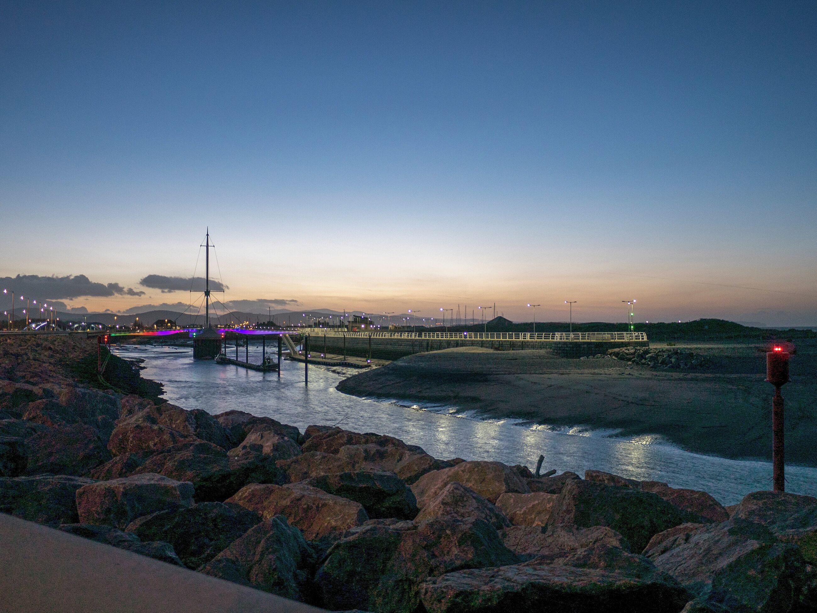 A coastal scene with a bridge, rocky shoreline, and a body of water reflecting lights.