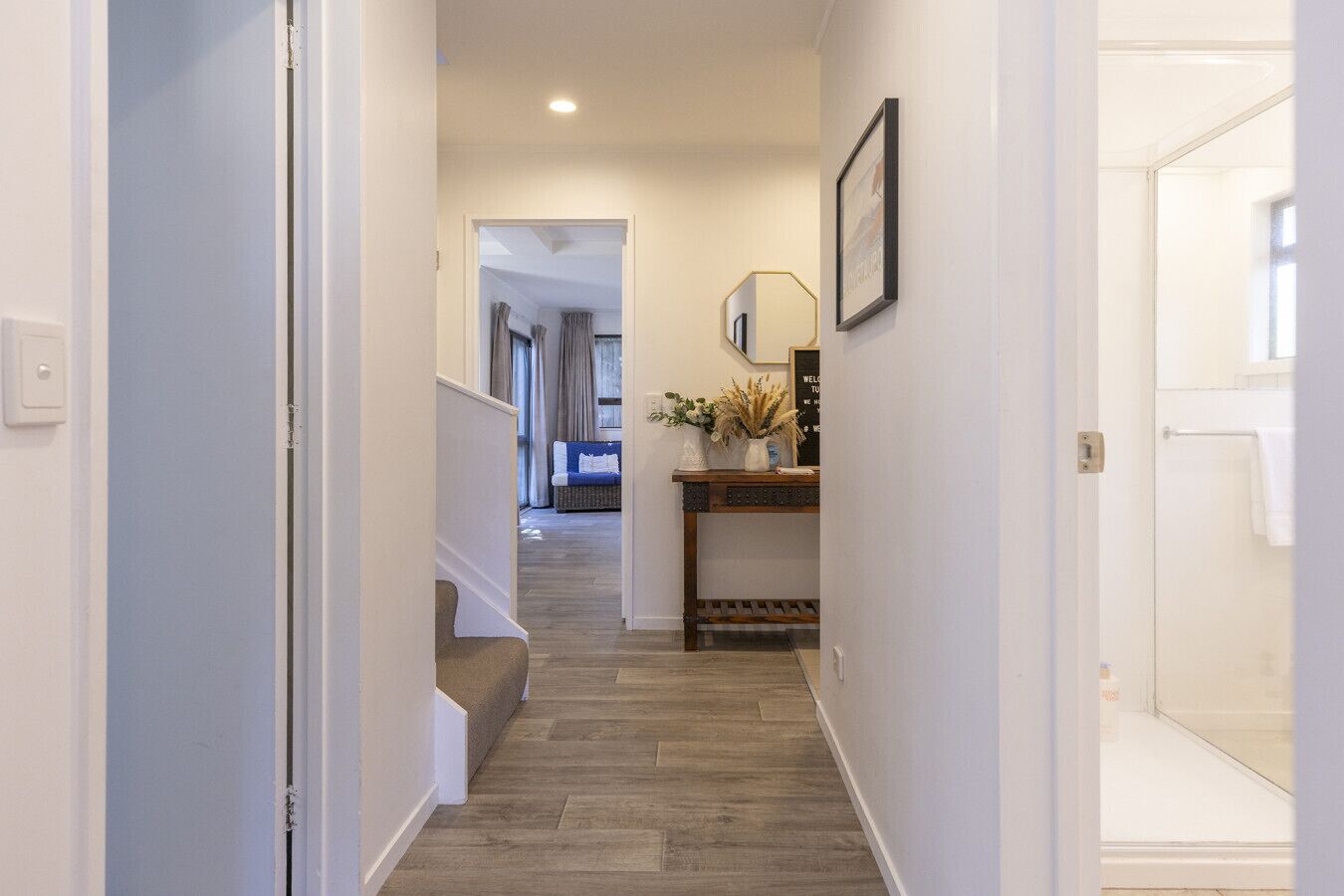 A hallway with a wooden floor, a staircase, a wall-mounted lamp, and a welcome sign.