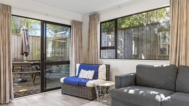 A cozy living room with a grey sofa, a wicker chair with a blue throw, and a small round table. Large windows offer views of a wooden deck and trees. - The Tui`s Nest - central to town (Taupō)