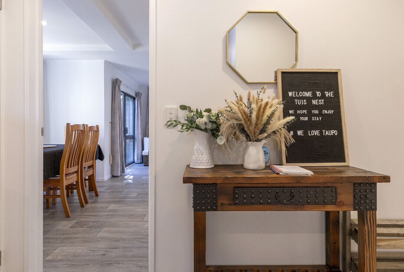 A reception area with a wooden table displaying a welcome sign, a vase with dried plants, and a mirror on the wall.