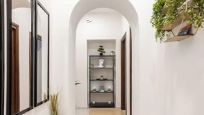 A hallway with a white wall, a wooden door, and a black-framed mirror. There is a plant on the right and a shelf with decorative items in the center. - Vatican Area | Stylish 2BR + Fast Wi-Fi & A/C (Roma)