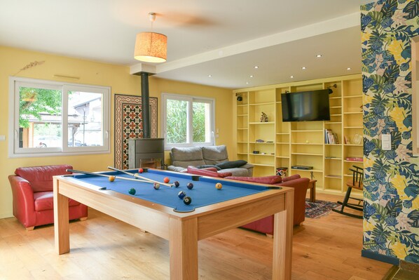 Une table de billard dans une pièce où se trouvent un canapé, un téléviseur posé sur une étagère et une fenêtre.