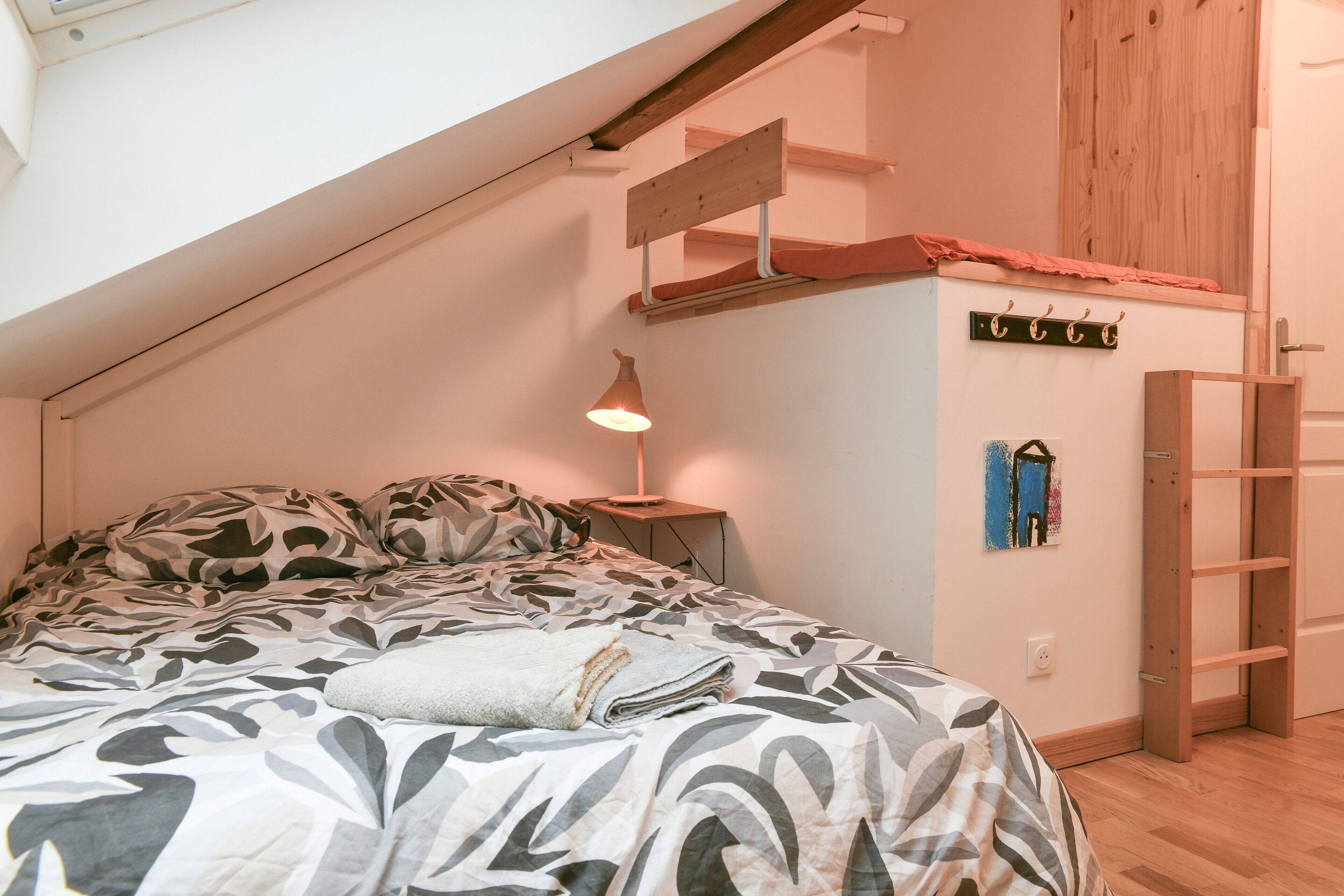 Une chambre avec un plafond en pente, un lit avec une literie à motifs, une lampe de chevet, une échelle en bois et un porte-manteau fixé au mur.