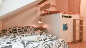 Une chambre avec un plafond en pente, un lit avec une literie à motifs, une lampe de chevet, une échelle en bois et un porte-manteau fixé au mur.