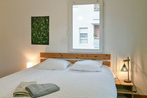 Un lit avec une literie blanche, une tête de lit en bois, une fenêtre donnant sur des immeubles, une œuvre d’art accrochée au mur et une lampe de chevet.