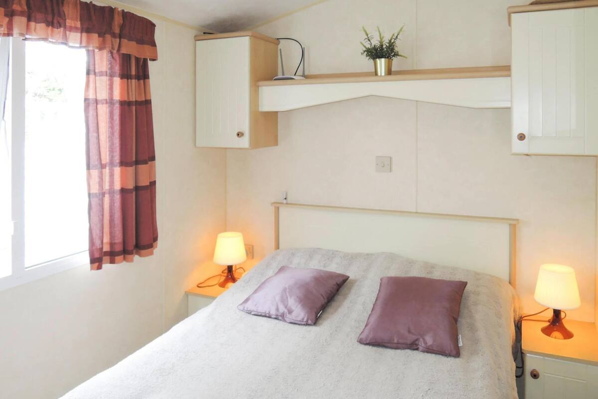 Une chambre  à coucher avec un lit, deux oreillers, des lampes de chevet, une fenêtre avec des rideaux et une étagère avec une plante.