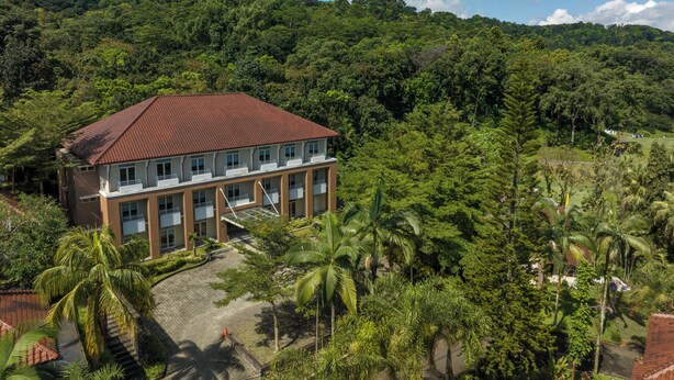 A multi-story building with a red-tiled roof, surrounded by lush greenery and palm trees. - Pelangi Hotel at Artotel Pelangi Park (Bogor)