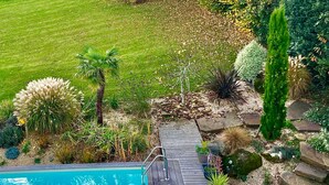 A swimming pool with a wooden deck, surrounded by various plants and a grassy lawn. - The Norman chalet (Les Champeaux)