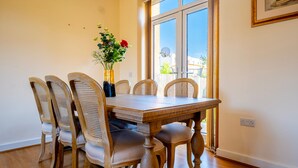 A dining area with a wooden table and six wicker chairs, a vase with flowers, and a window with a view of the outside. - 14 Guests-6 Bedroom House, Free Parking (Milton Keynes)
