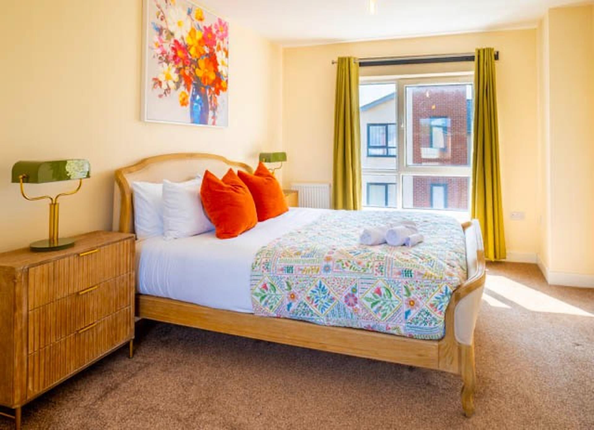 A bedroom with a wooden bed, white bedding, orange pillows, a colorful quilt, a green lamp, and a window with yellow curtains.