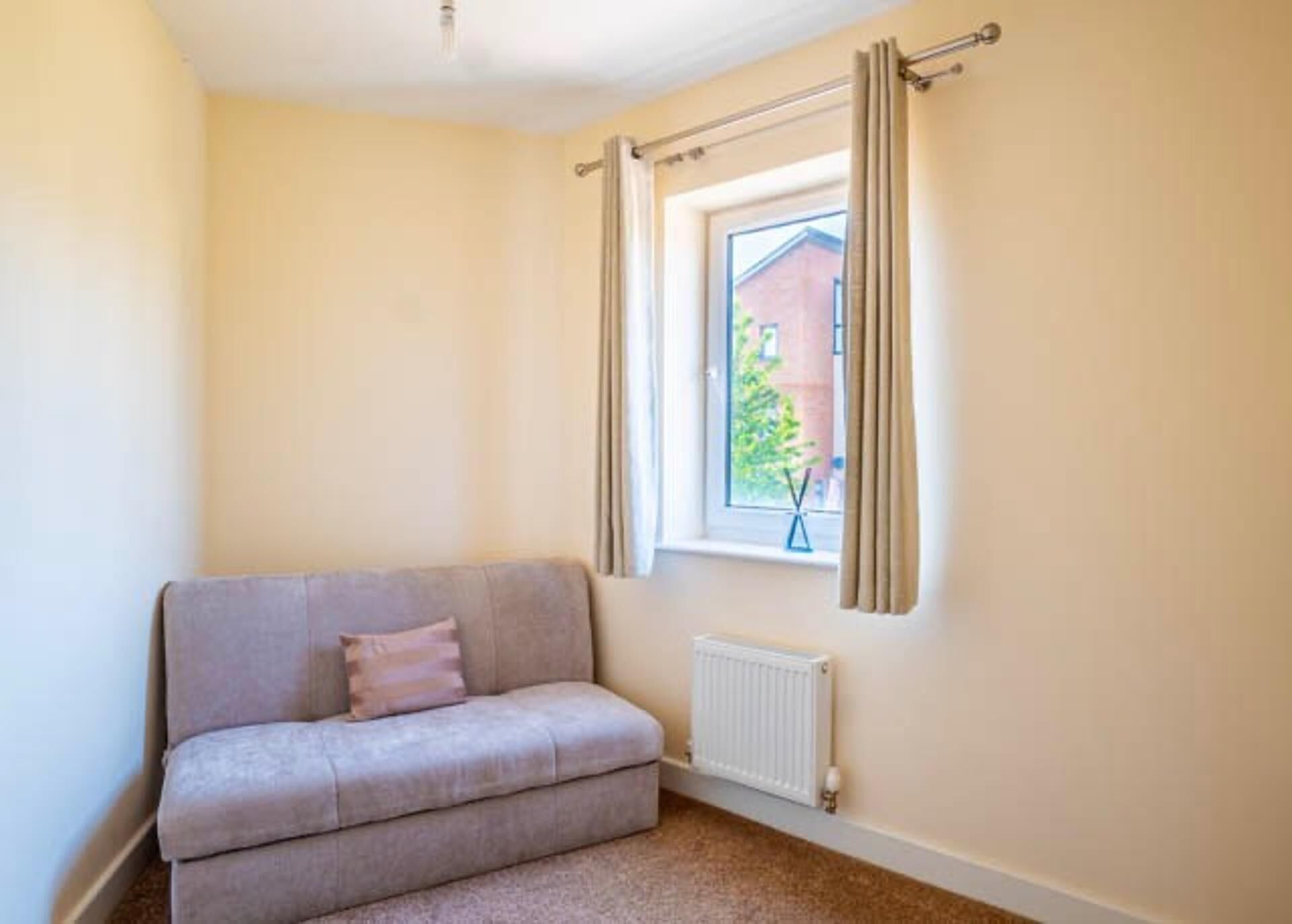 A small, cozy room with a grey sofa, a window with curtains, and a radiator.