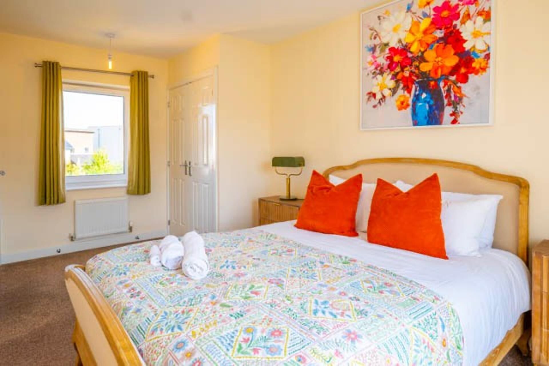 Un dormitorio con cama, dos almohadas, una mesita de noche, una lámpara, una ventana con cortinas y un cuadro en la pared.