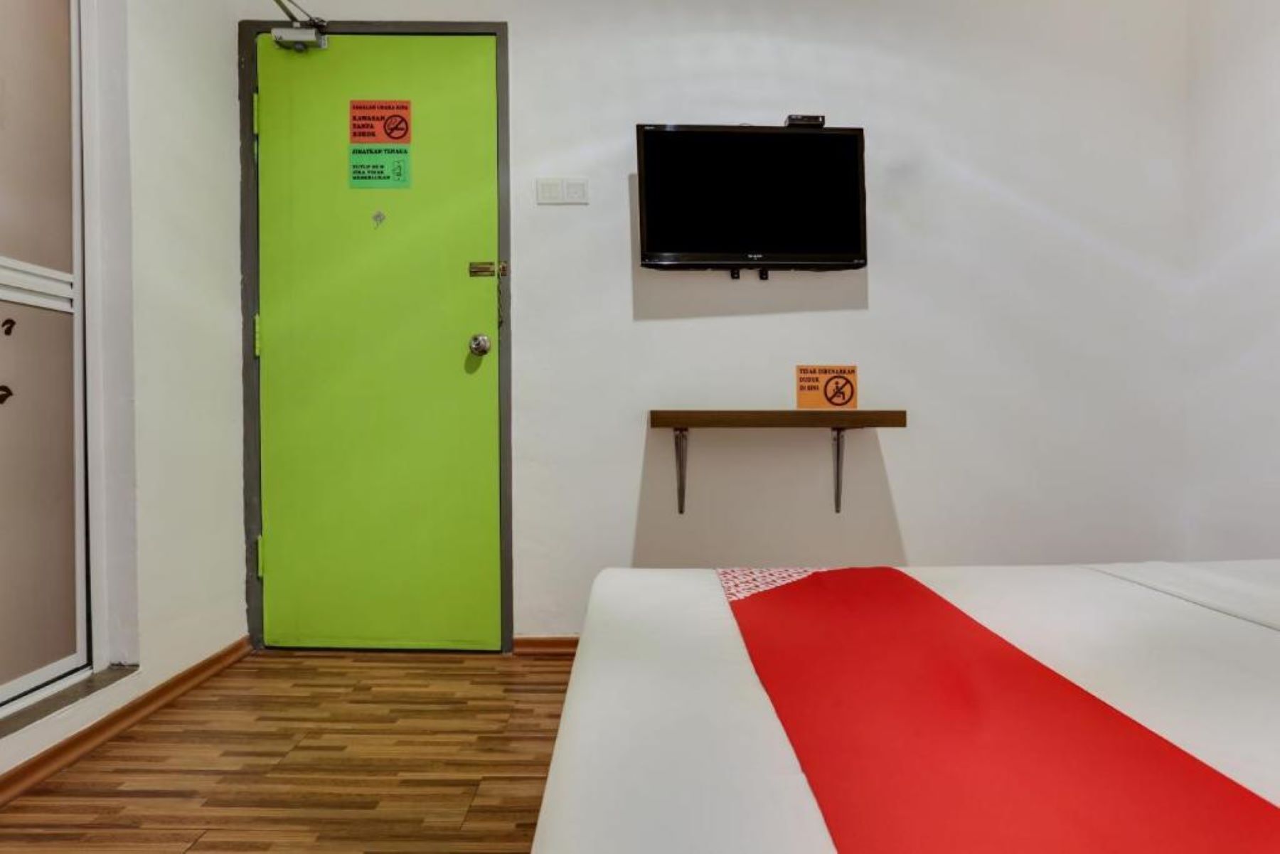 A hotel room with a green door, a bed with a red and white bedspread, a mounted television, and a small shelf with a sign.