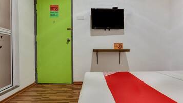 Una habitación de hotel con una puerta verde, una cama con colcha roja y blanca, un televisor montado y un estante con un letrero.