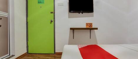 A hotel room with a green door, a bed with a red and white bedspread, a mounted television, and a small shelf with a sign.