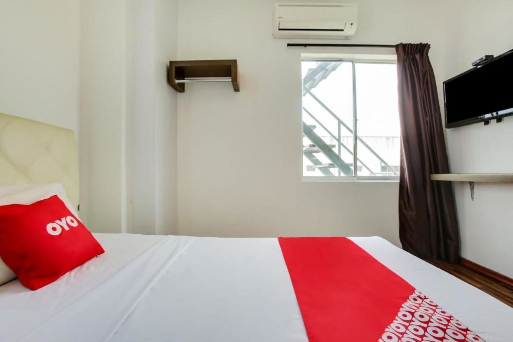 Quarto de hotel com cama, janela com vista para uma escadaria, ar condicionado e televisão na parede.