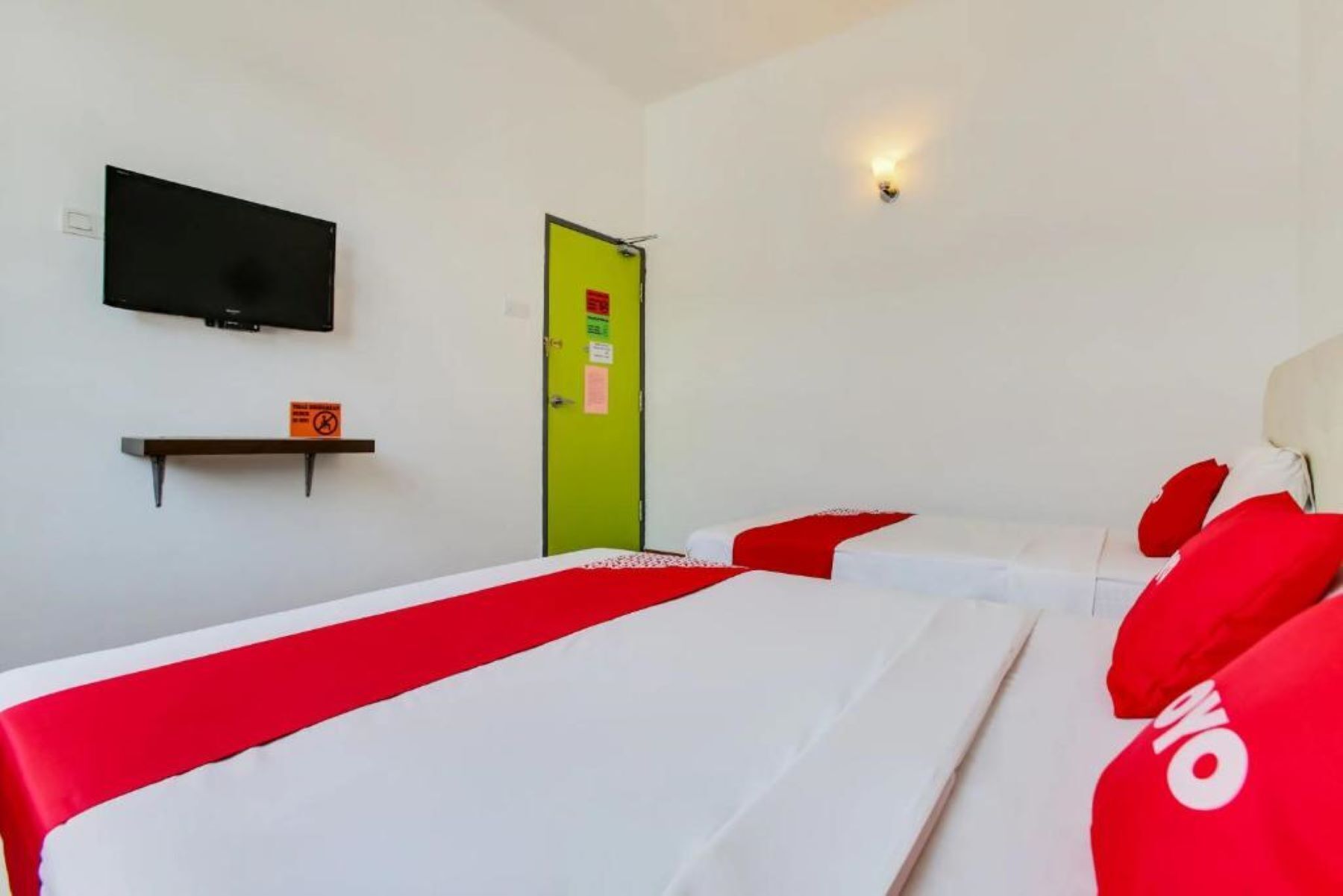 A hotel room with two beds, a television on the wall, a green door, and a shelf with a sign.