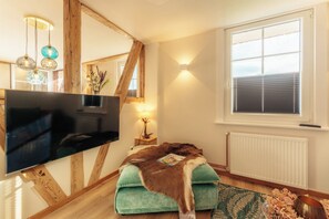 A modern living room with a large flat-screen TV, wooden beams, a green cushioned bench, a small side table with a lamp, and a window with blinds.