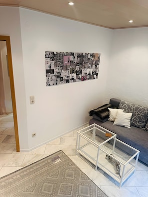 Un salon comprenant un canapé, une table basse en verre, un tapis à motifs et un mur décoré d’un collage artistique.