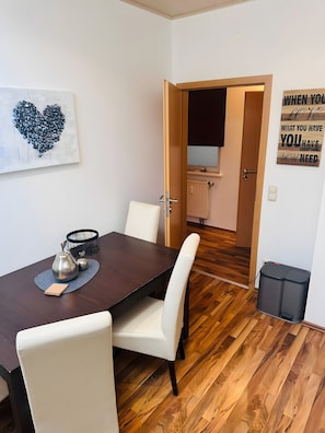 Un coin repas avec une table en bois, des chaises blanches, une œuvre d’art en forme de cœur et une citation inspirante sur le mur.