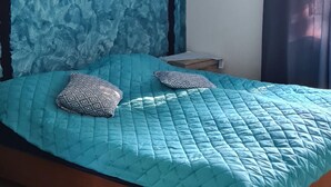 Una camera da letto con una coperta trapuntata blu, due cuscini decorati, una testiera in legno, una ventola a soffitto e una finestra con tende.