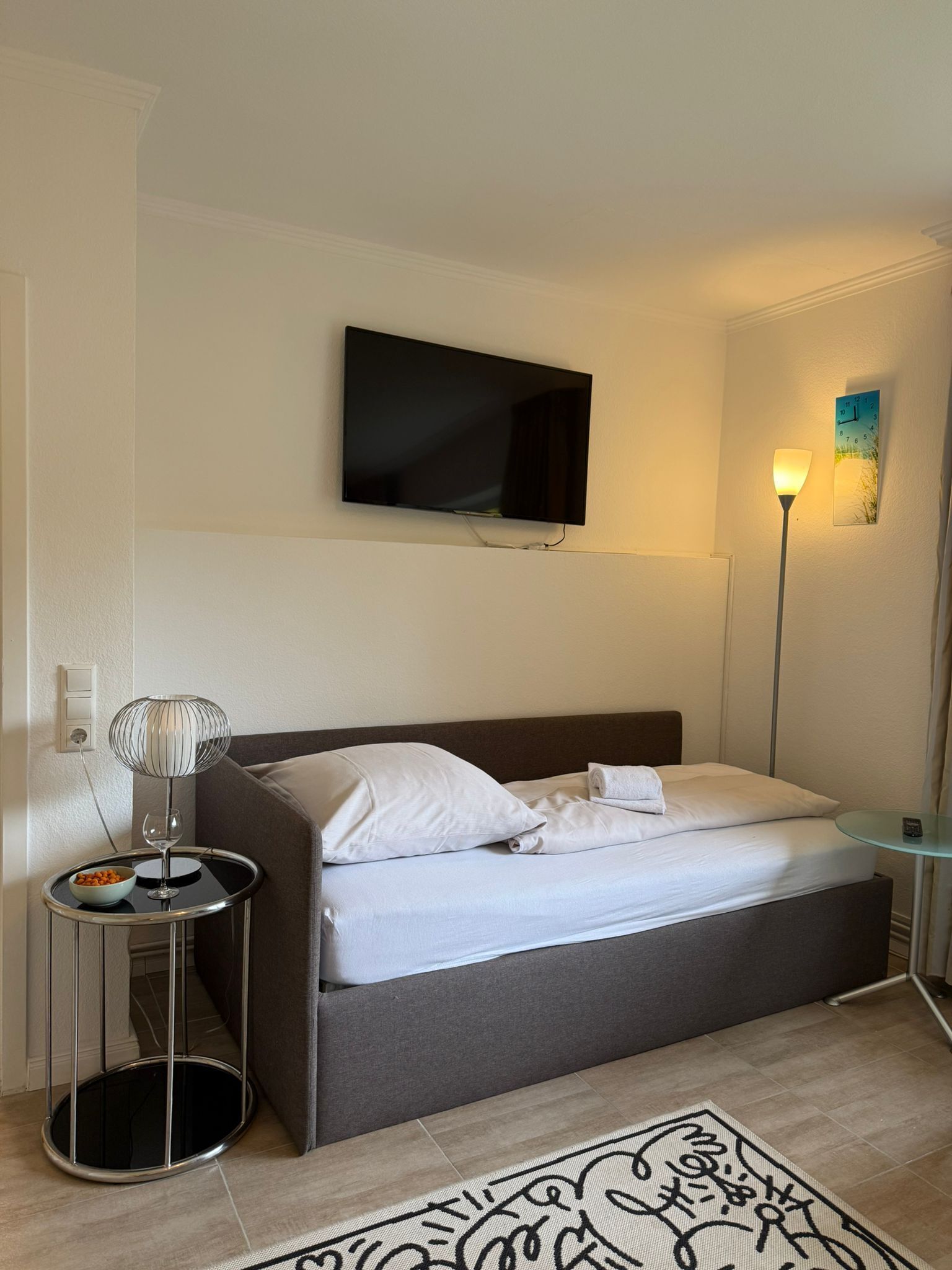 A hotel room with a bed, a TV mounted on the wall, a bedside lamp, a small table with a bowl and a glass, and a patterned rug on the floor.