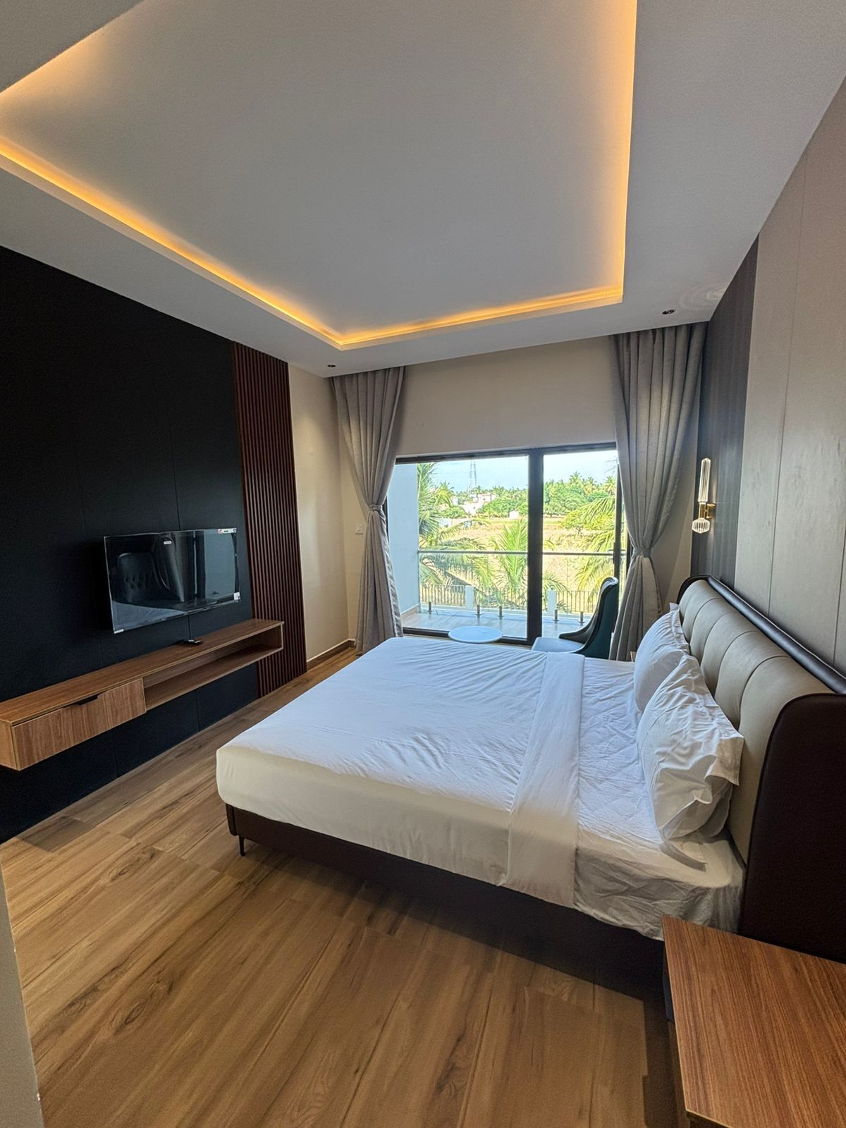 Ein modernes Schlafzimmer mit einem großen Bett, einem an der Wand montierten Fernseher und einem Balkon mit Blick auf Grünflächen.