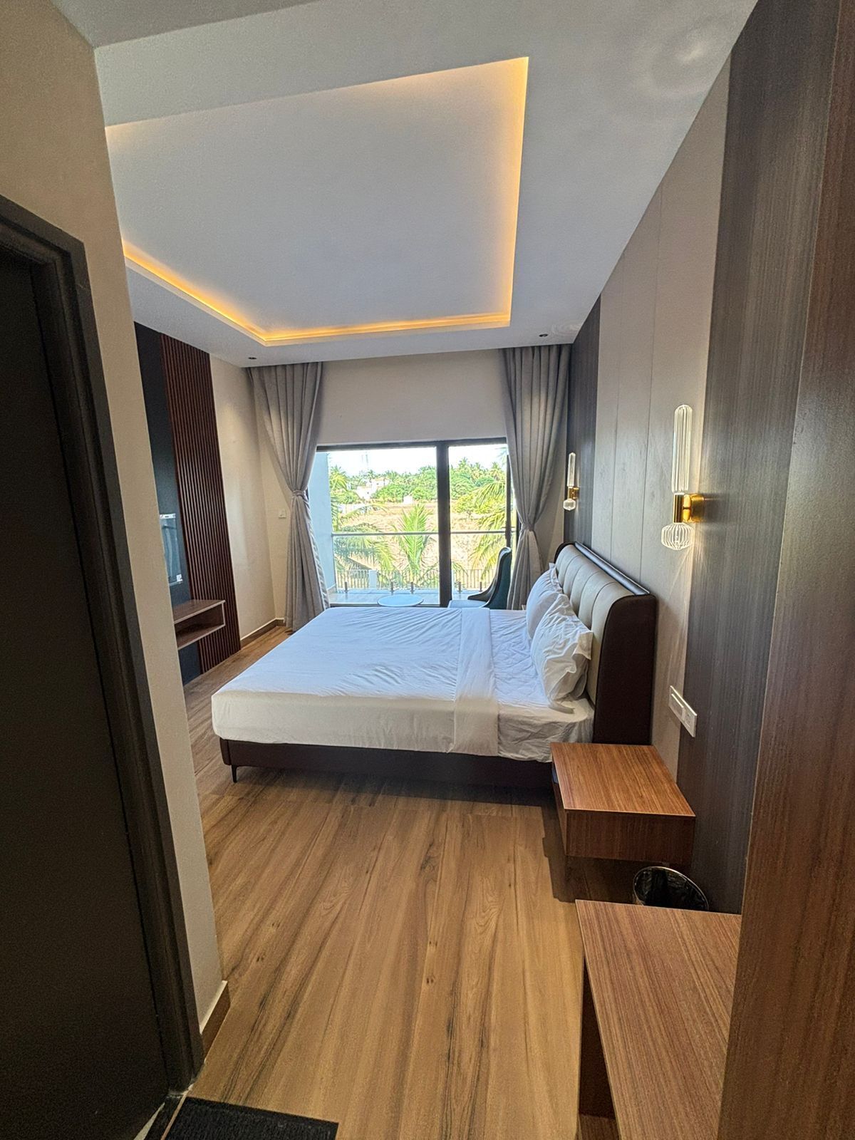 A modern bedroom with a large bed, wooden flooring, and a view of greenery outside.