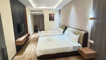 A modern hotel room with two beds, wooden flooring, a flat-screen TV, a wall-mounted light, and a small table.