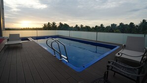 En pool på taket med ett trädäck, solstolar och utsikt över palmer och berg i bakgrunden.