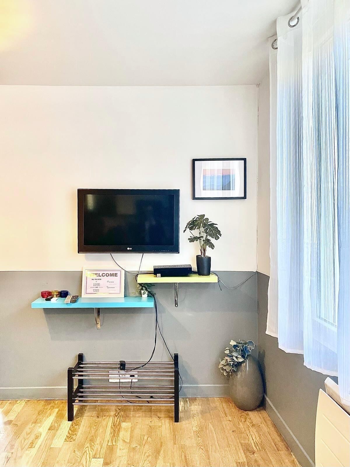 Ein Wohnzimmer mit einem an der Wand befestigten Flachbildfernseher, einem kleinen Regal mit einer Pflanze, einem gerahmten Bild und einem Heizkörper.
