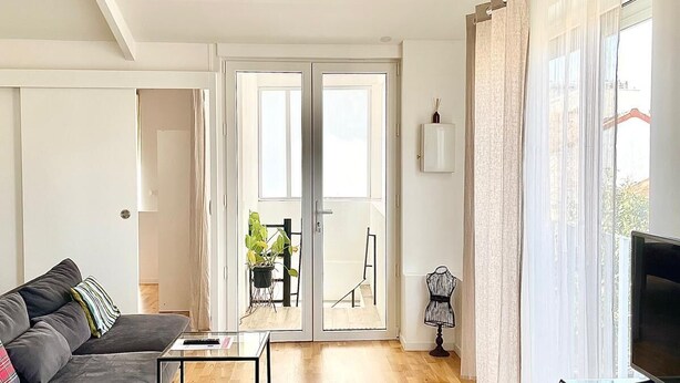 Ein modernes Wohnzimmer mit einer Couch, einem Hocker und einem Glastisch. Ein Fernseher steht auf einem Schrank und eine Topfpflanze ist ebenfalls vorhanden.
