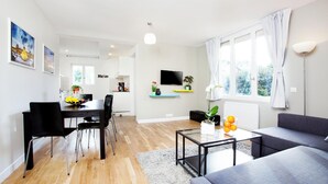 Ein modernes Wohnzimmer mit einem grauen Sofa, einem Esstisch mit Stühlen, einem Couchtisch mit Pflanze und Orangen, einem Fernseher an der Wand und einem Fenster mit Vorhängen.