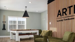 A modern living room with a pool table, green armchairs, and a large window with blinds.