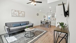 A modern living room with a grey sofa, a coffee table, a ceiling fan, a dining area with chairs, and a kitchen in the background. - Arte Luxury townhomes O5 I Comfy 2 Bed 2.5 Bath (Clarksville)